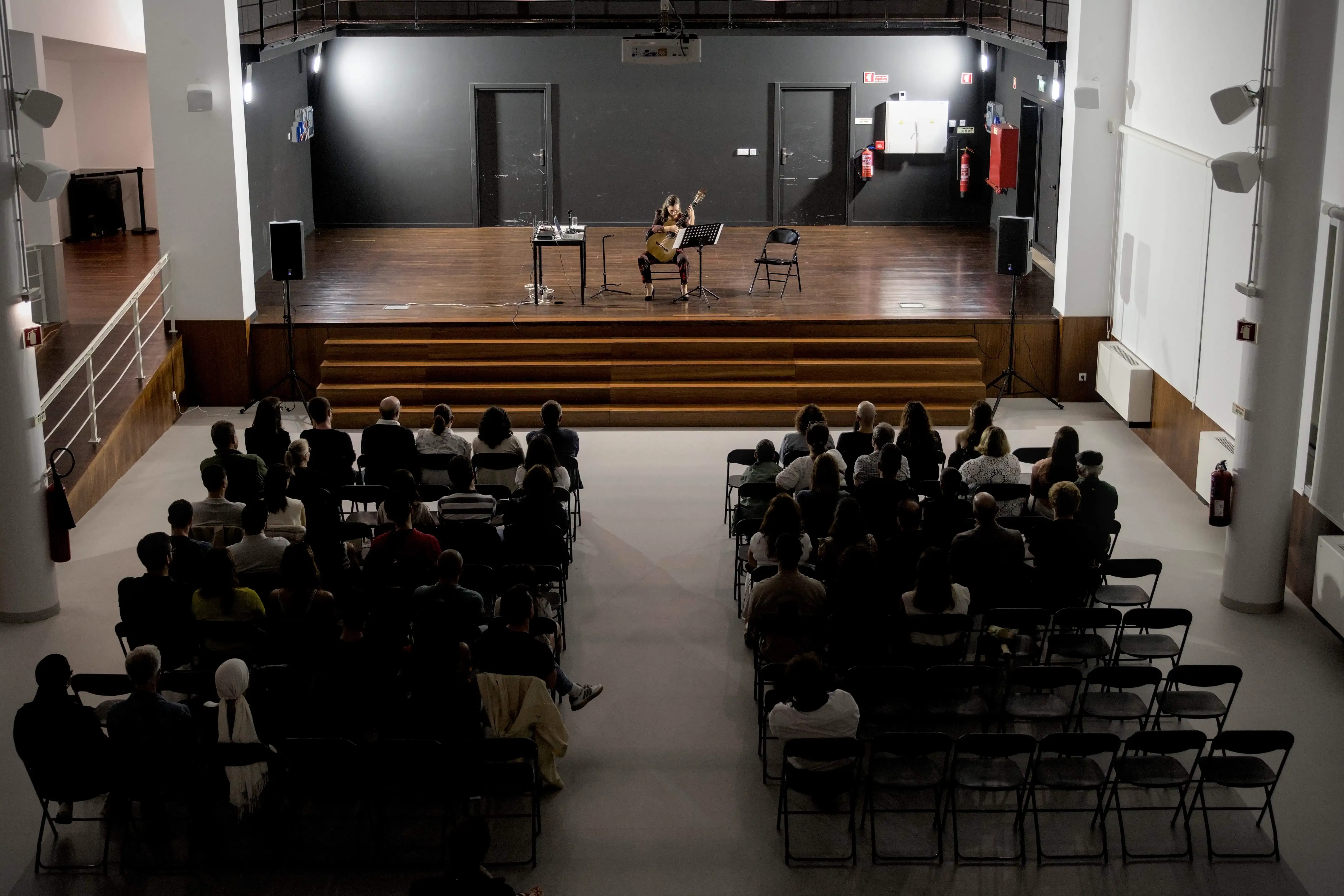 Uma mulher toca guitarra clássica num palco. Perspetiva superior revelando toda a plateia